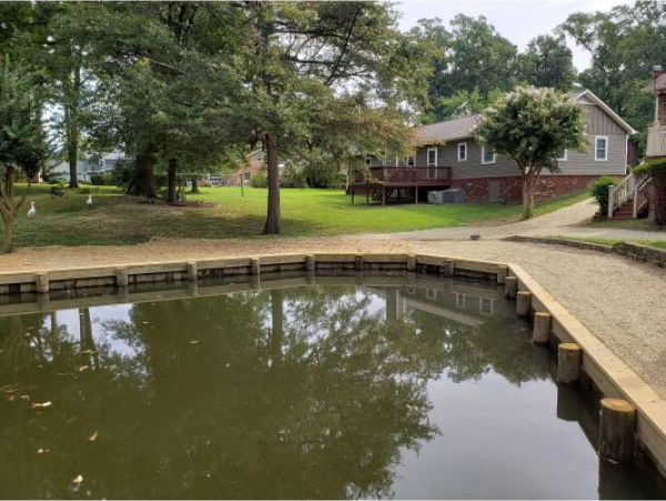 Pond with wood border, gravel path, and house in background. Reflective water surface.