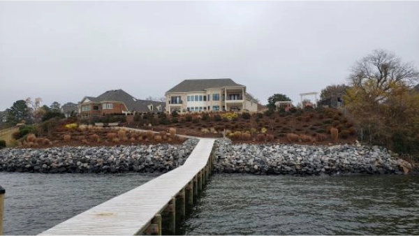 Wooden dock leading to a large house on a rocky shore, under a cloudy sky.