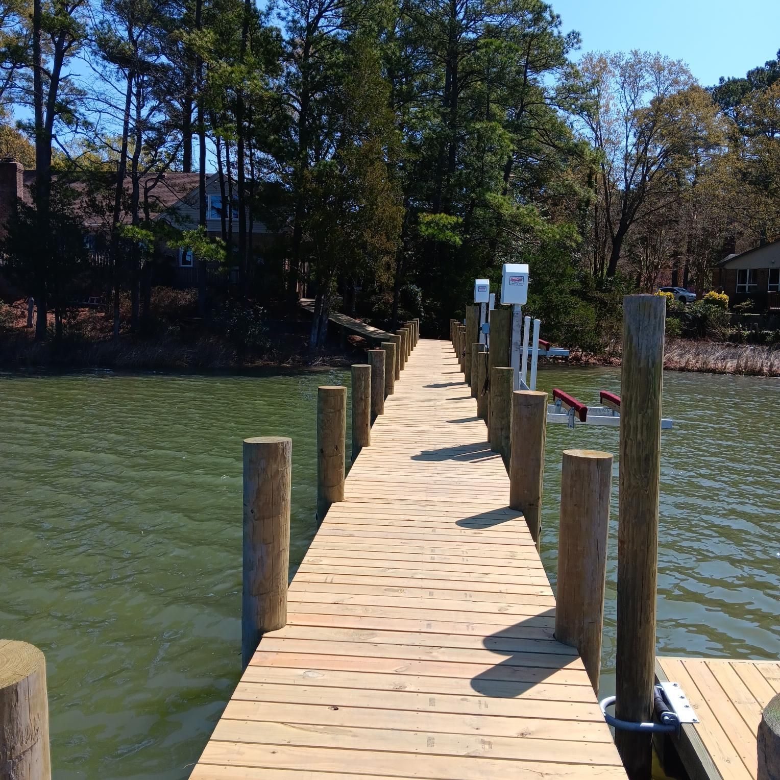 Wooden pier extending into green water, surrounded by wooden posts. Houses and trees in the background. Sunny day.