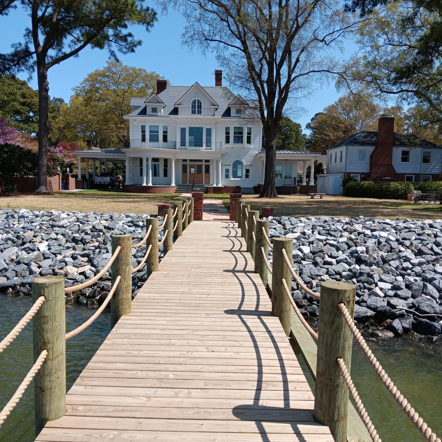 Wooden dock leads to white house on a sunny day. Large trees and a rocky shoreline.