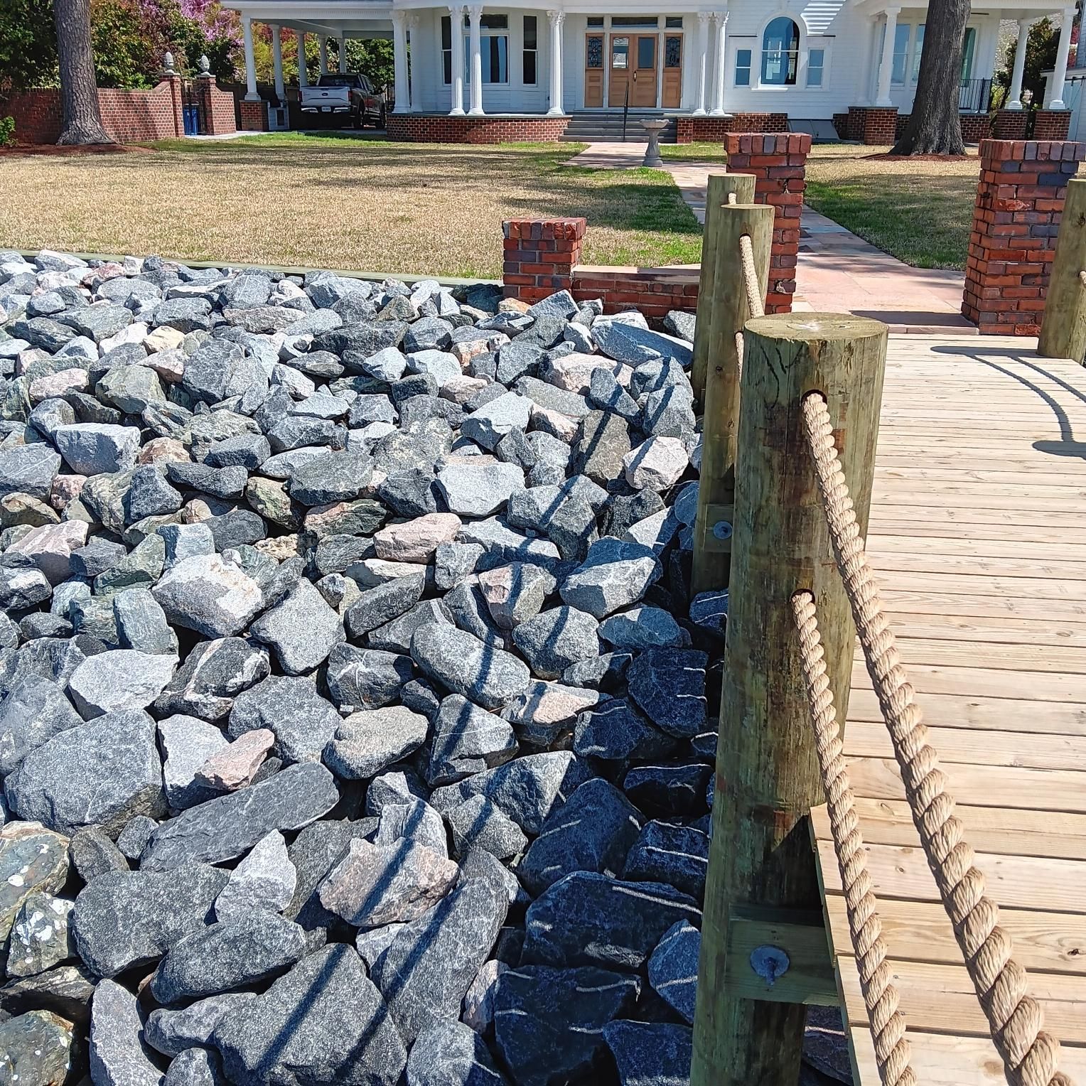Rocks lining a shore next to a wooden pier, with a white house in the background.