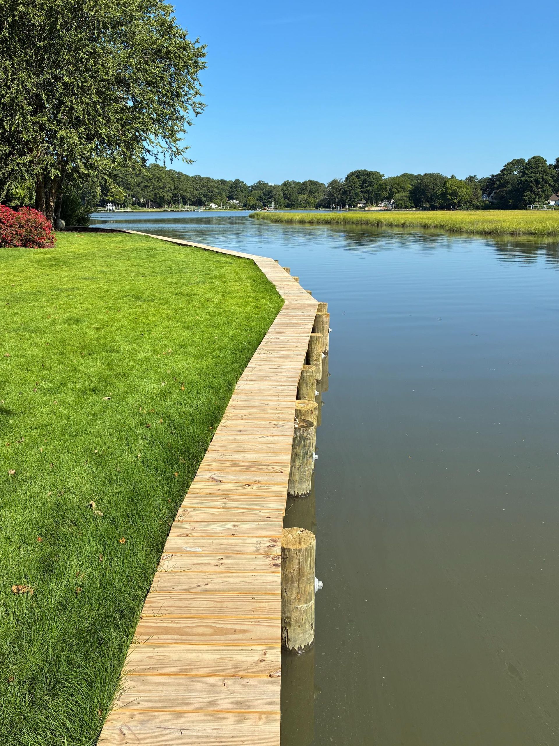 Wooden walkway along a body of water with green grass and trees on a sunny day.