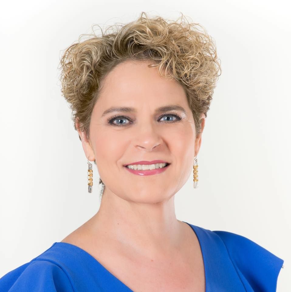 A person with short, curly blonde hair and blue eyes, smiling, wearing a blue top and gold earrings against a white background.