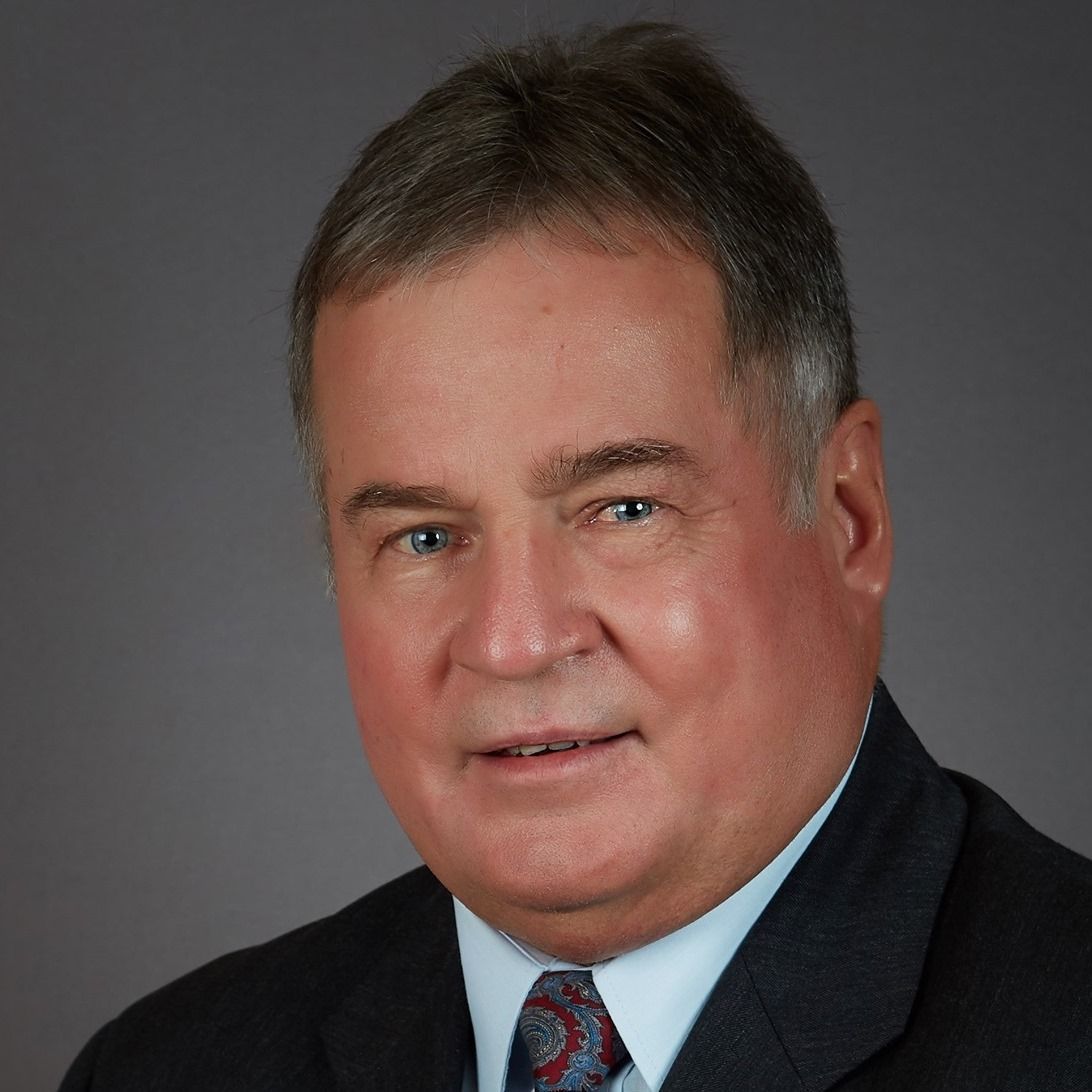 A professional headshot of a man wearing a dark blazer, light blue shirt, and patterned tie against a grey background.