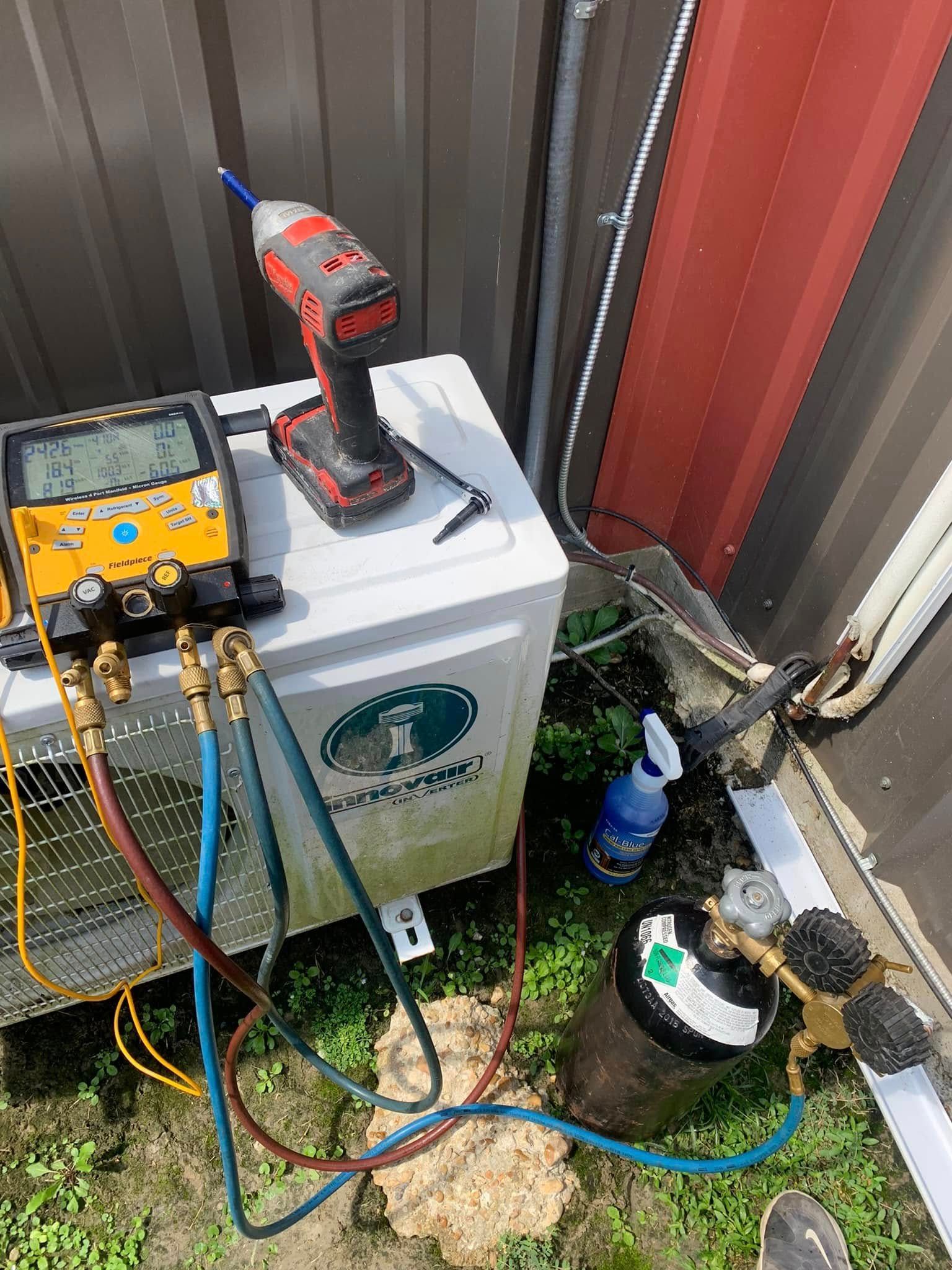 HVAC digital gauges and an impact driver sit on an outdoor unit with refrigerant hoses connected to a cylinder.