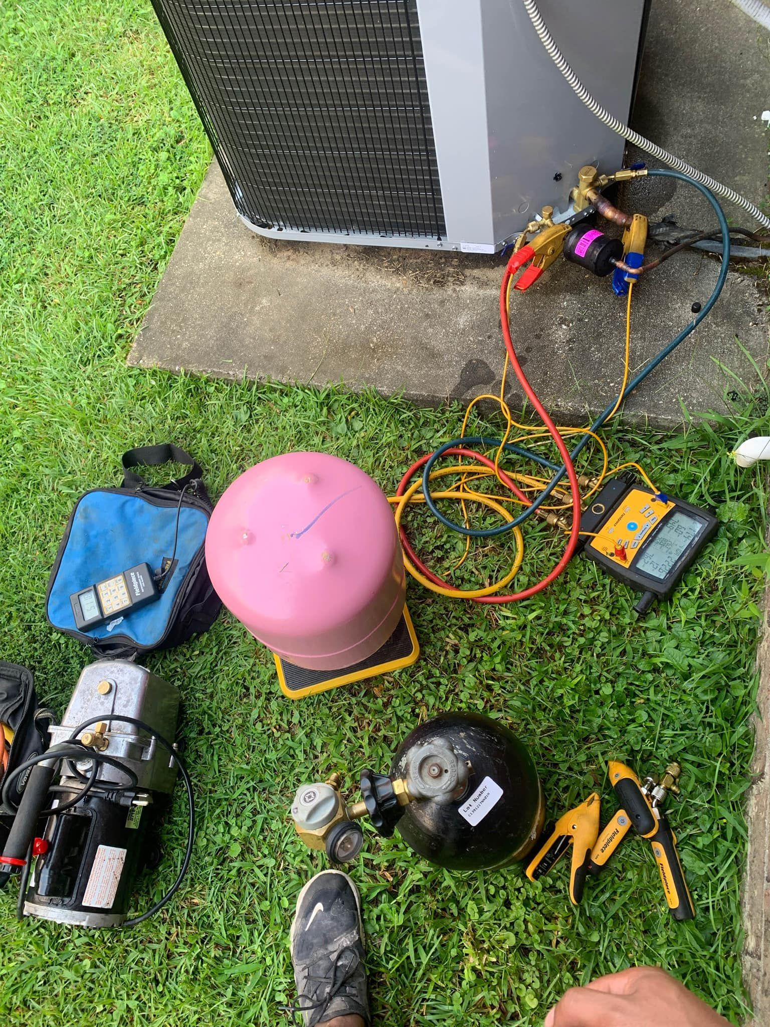 HVAC tools, including refrigerant tanks, a pump, and manifold gauges, arranged on grass beside an outdoor AC unit.