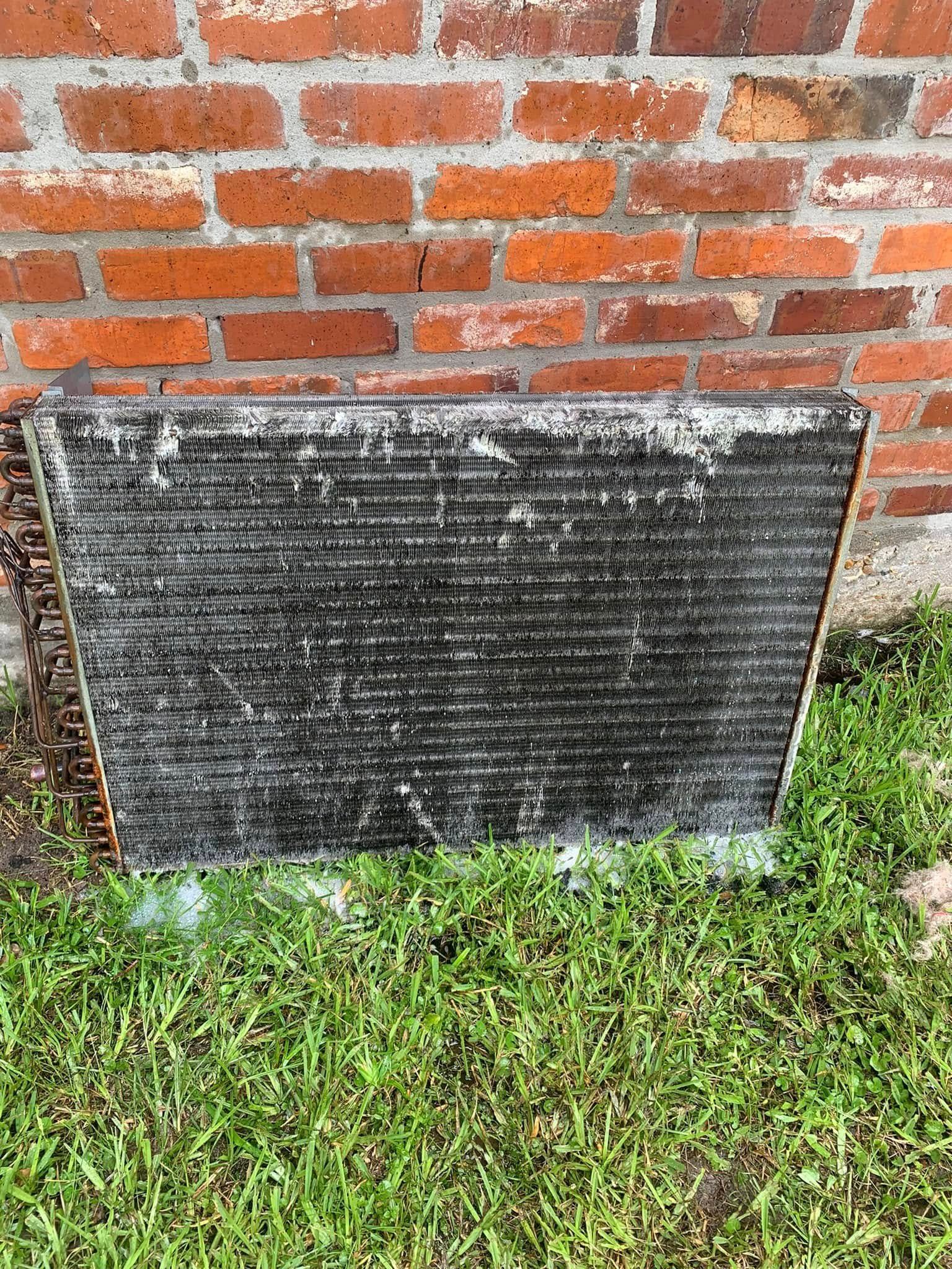 A dirty, rectangular HVAC radiator coil stands on green grass in front of a red brick wall.