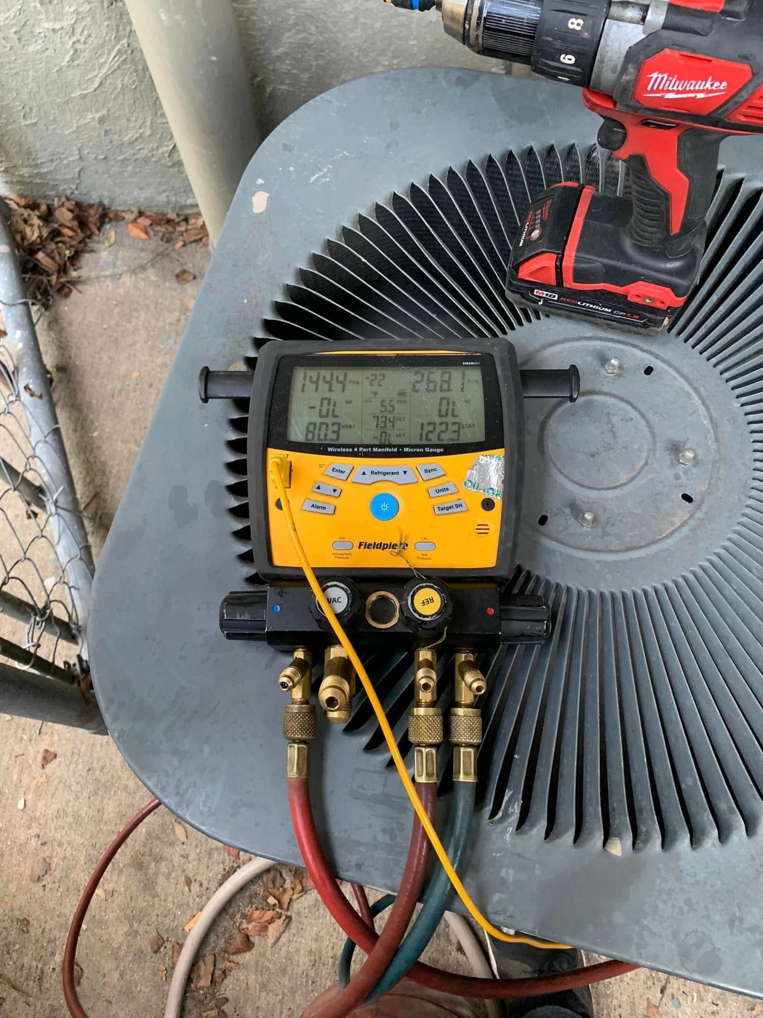 A yellow digital HVAC manifold gauge set connected to an outdoor air conditioning unit with a power drill nearby.