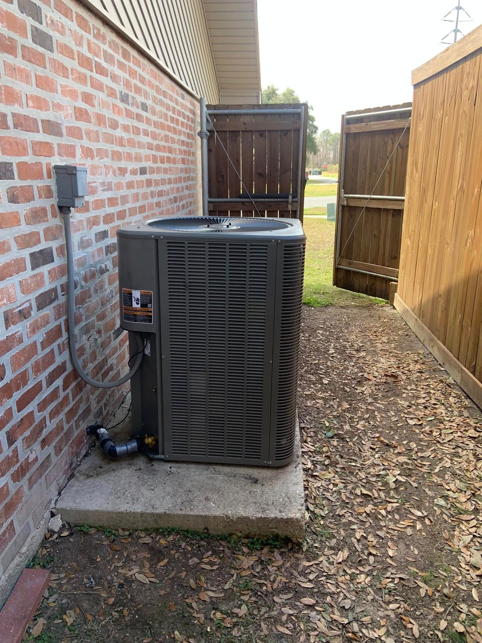 An outdoor HVAC unit sitting on a concrete pad next to a brick house wall, with a wooden fence in the background.
