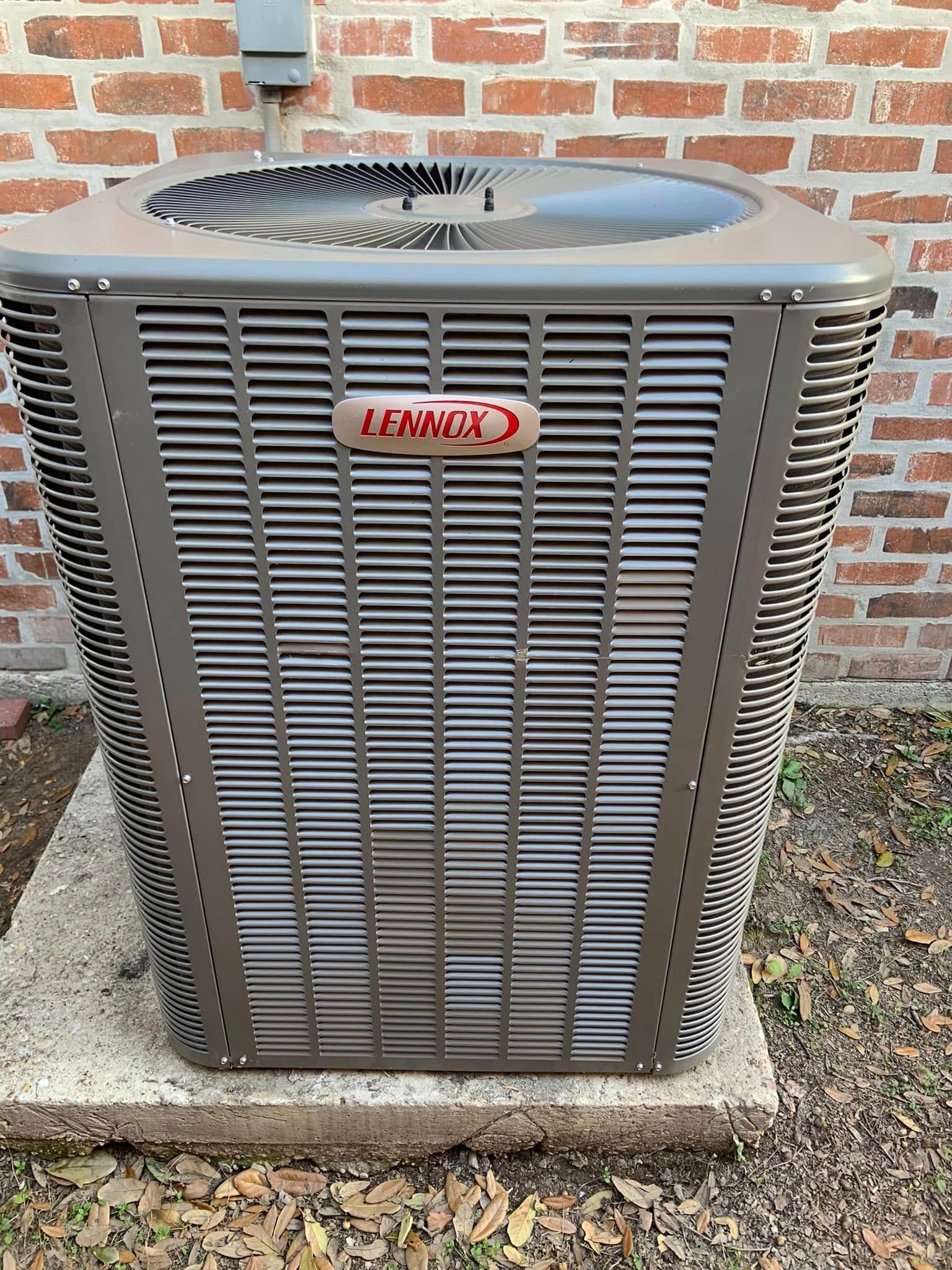 A Lennox central air conditioning unit sits on a concrete pad against a red brick exterior wall.