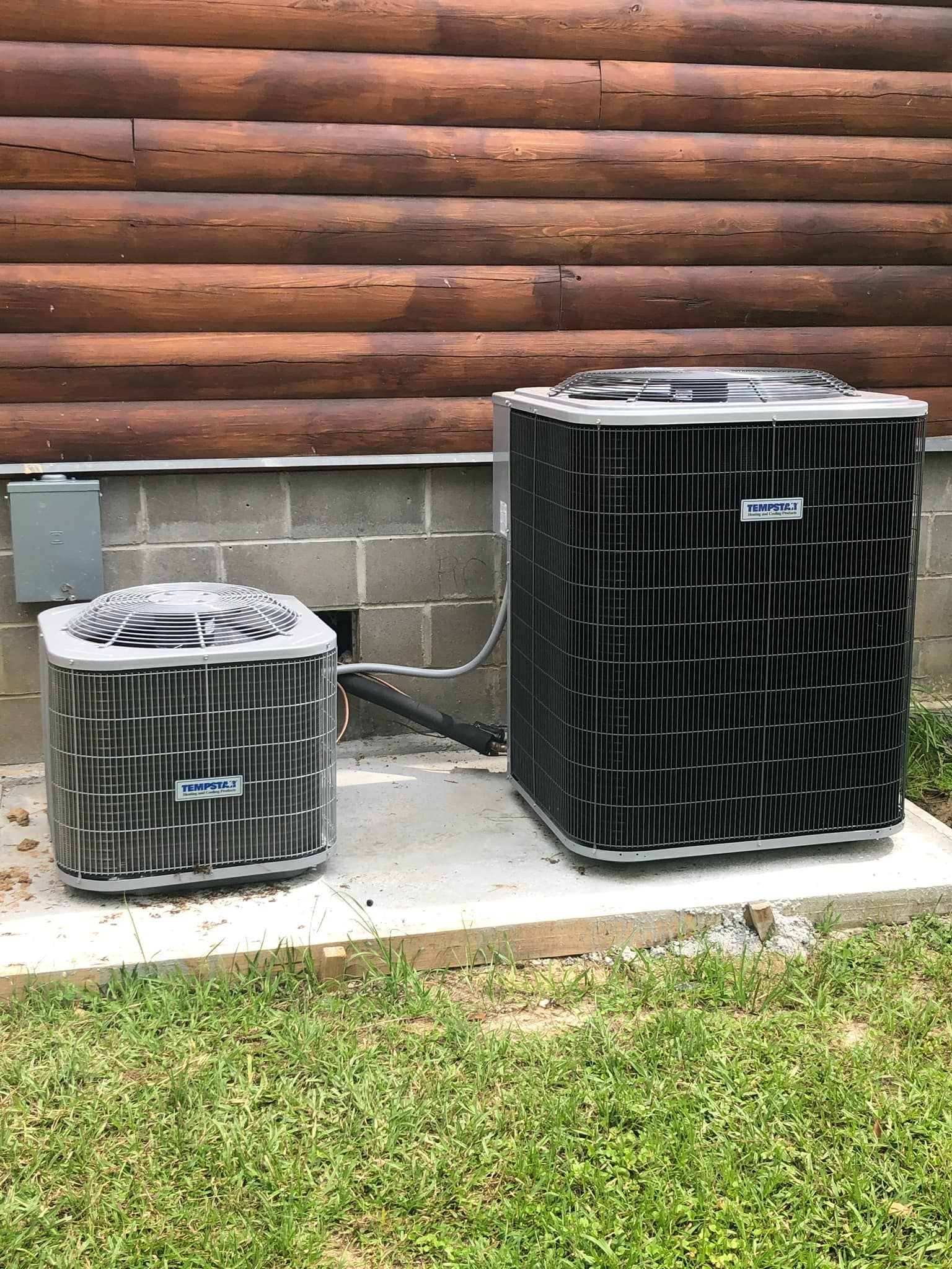 Two gray HVAC condenser units sit on a concrete slab next to a log cabin wall.