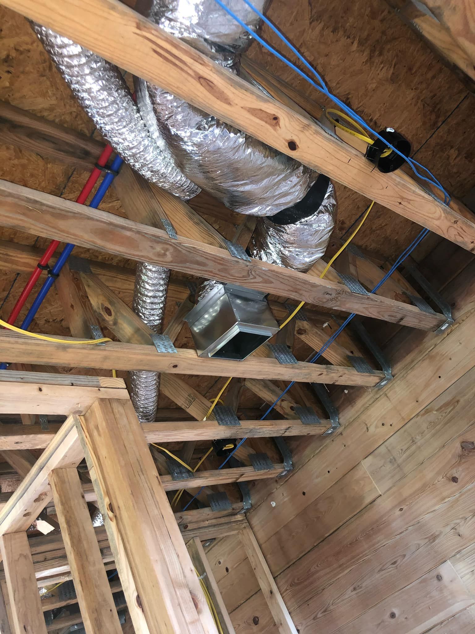 An unfinished ceiling frame with metal HVAC ducts, red and blue plumbing pipes, and yellow electrical wiring.