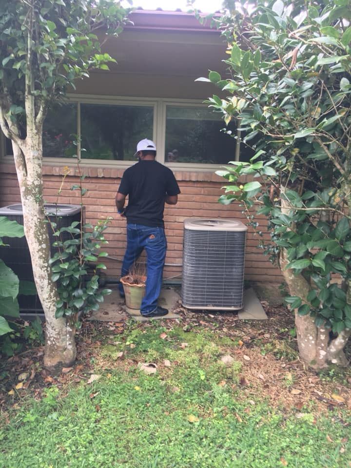 A person in a black shirt and blue jeans stands in a backyard near an air conditioning unit and a small tree.