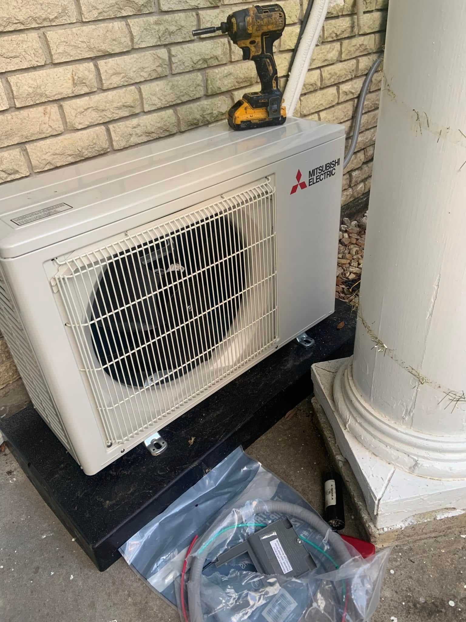 A Mitsubishi outdoor mini-split unit sits on a black mounting base against a brick wall next to a white column.