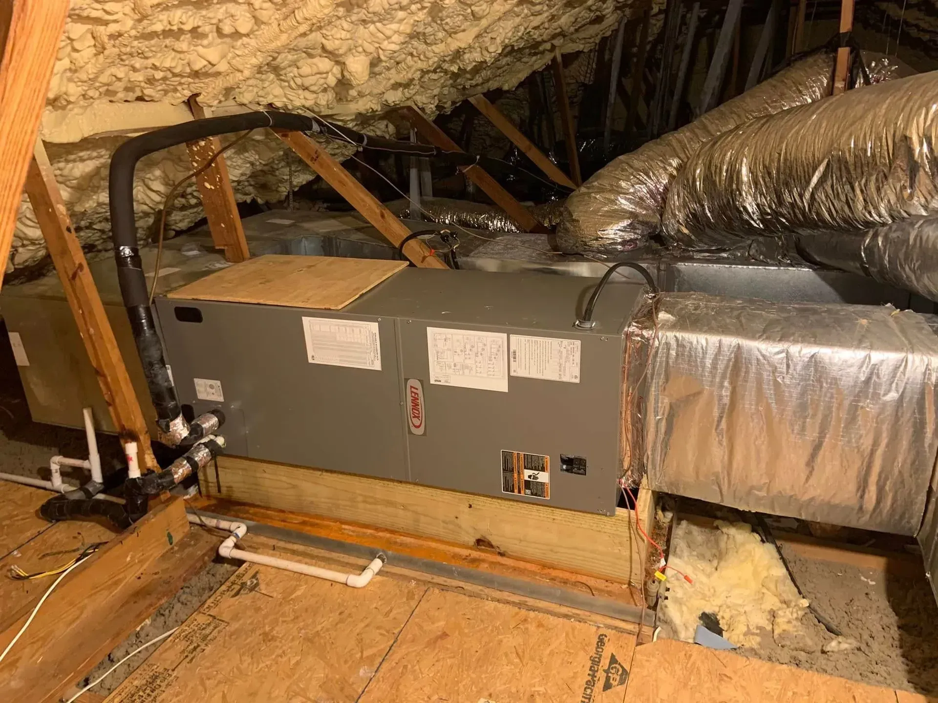 An HVAC air handler unit installed in an attic with insulated ductwork, spray foam roof insulation, and a wooden base.