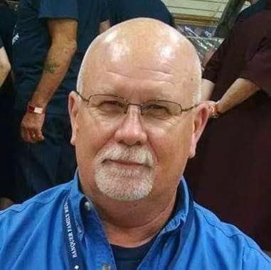 A person with a shaved head and glasses smiles slightly, wearing a blue collared shirt.