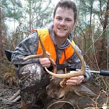 A person in camouflage hunting gear and a bright orange vest holds the antlers of a deer in a wooded outdoor setting.