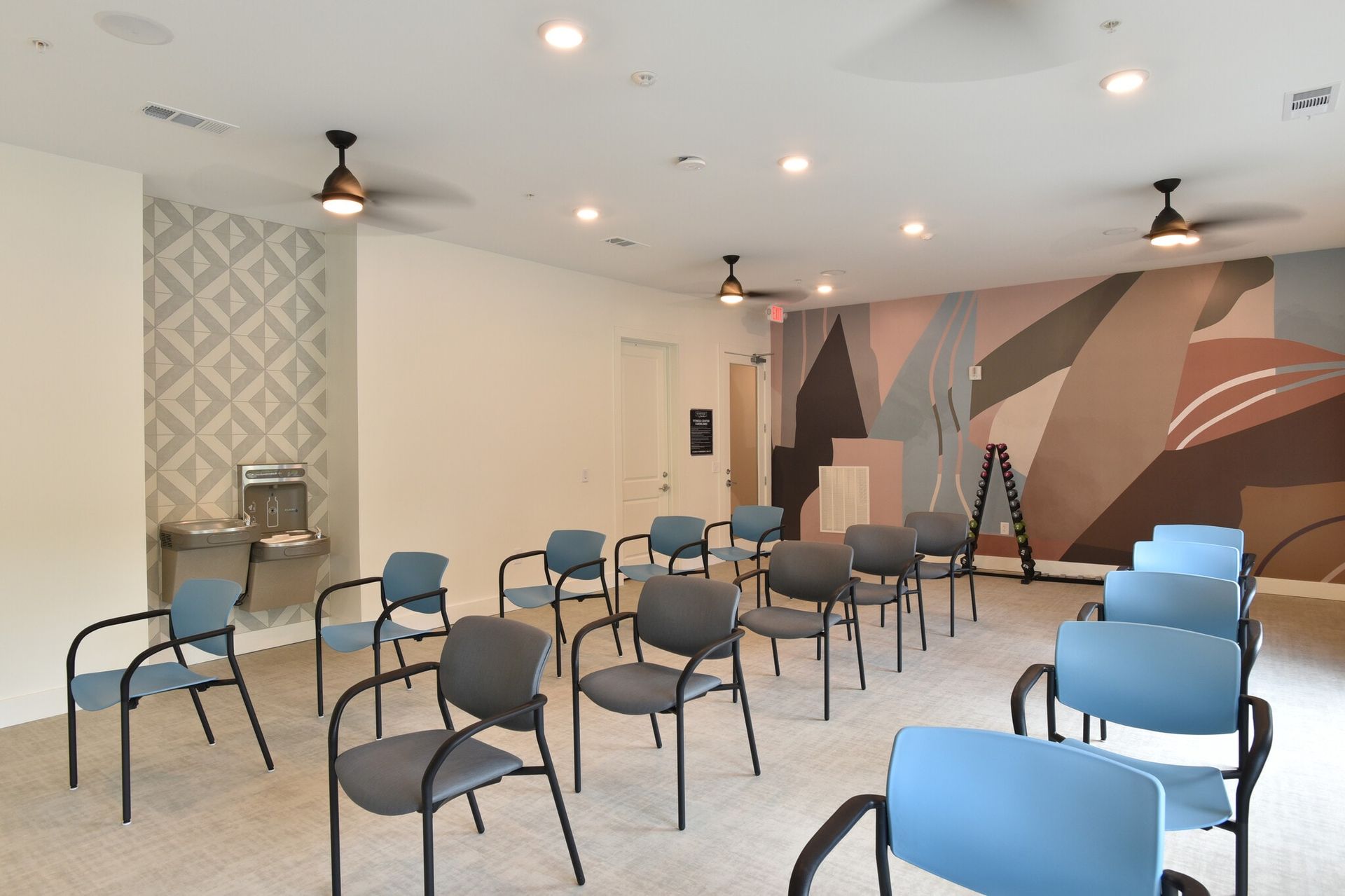 Bright meeting room with rows of blue and beige chairs, wall mirror, ceiling fans, and large windows