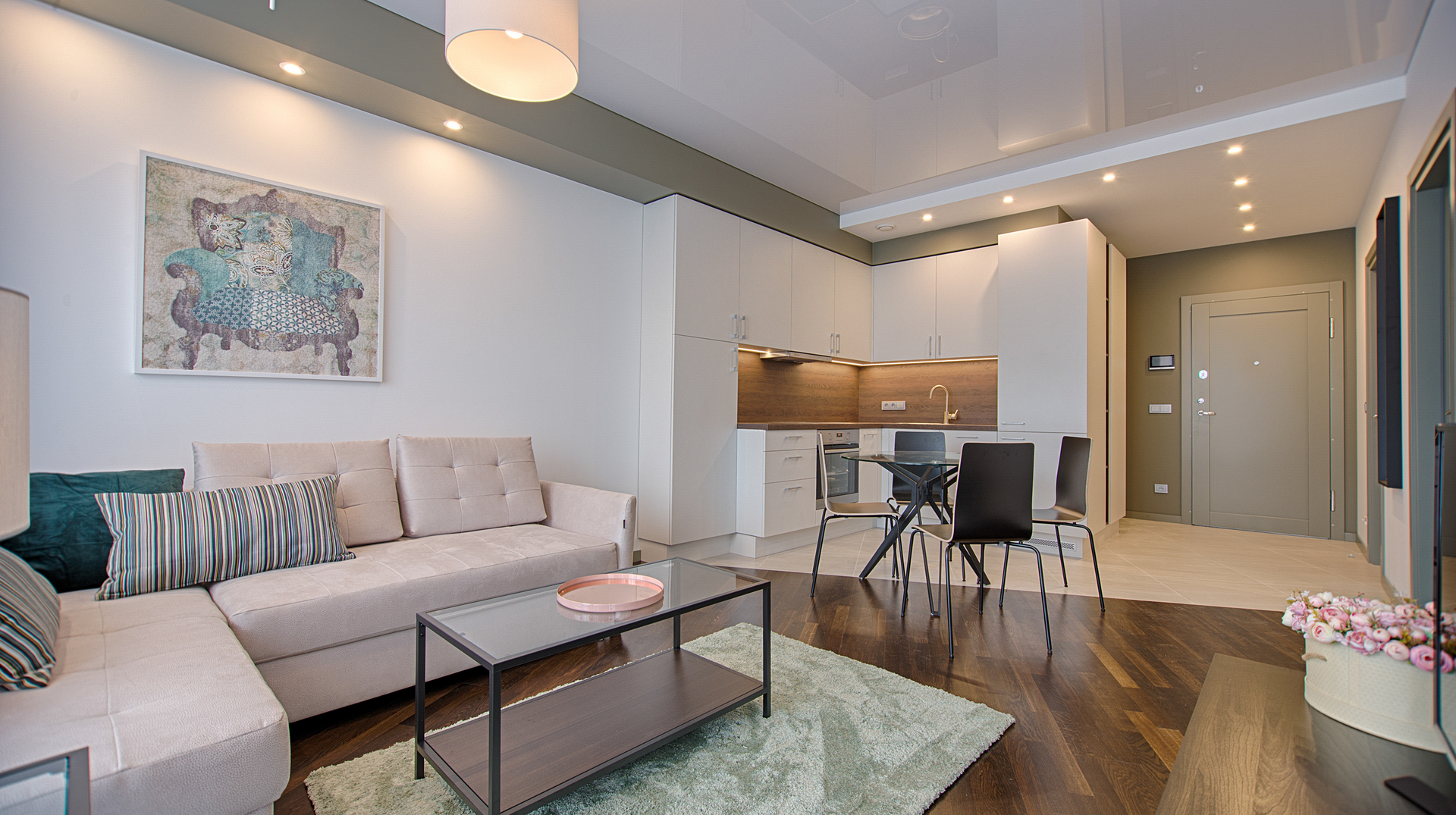Modern living room with beige sectional sofa, glass coffee table, dining area, and kitchen in the background.