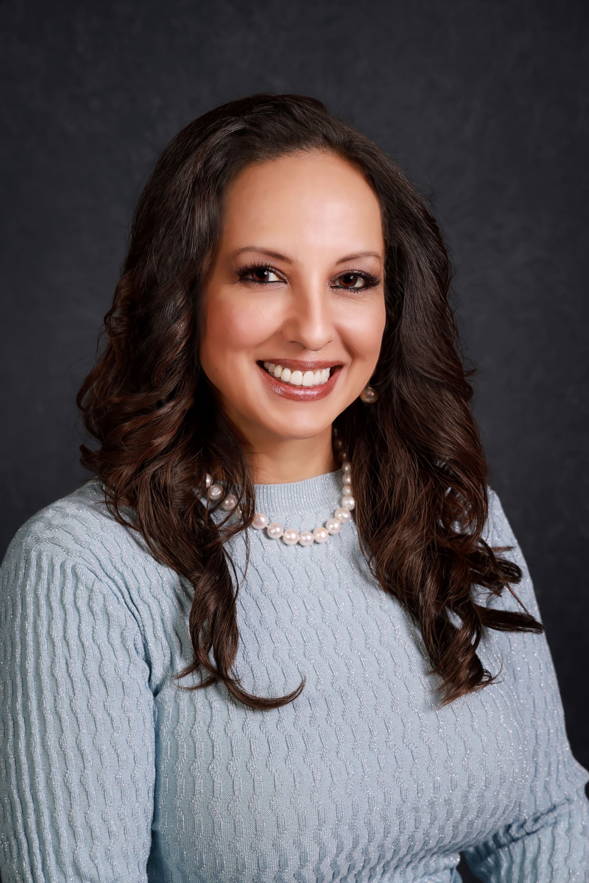A woman wearing a blue sweater and a pearl necklace is smiling for the camera A woman wearing a blue sweater and a pearl necklace is smiling for the camera