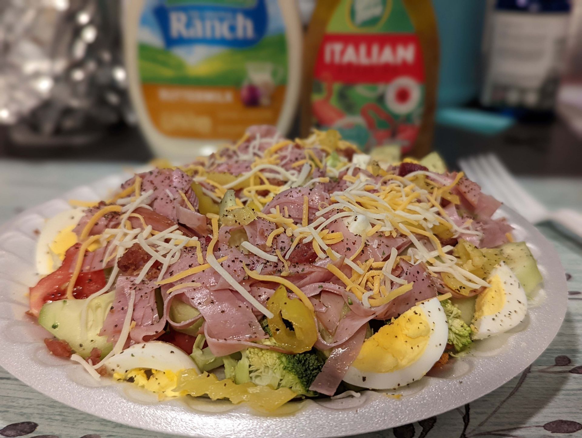 A paper plate topped with a salad with ham , eggs , broccoli and cheese.