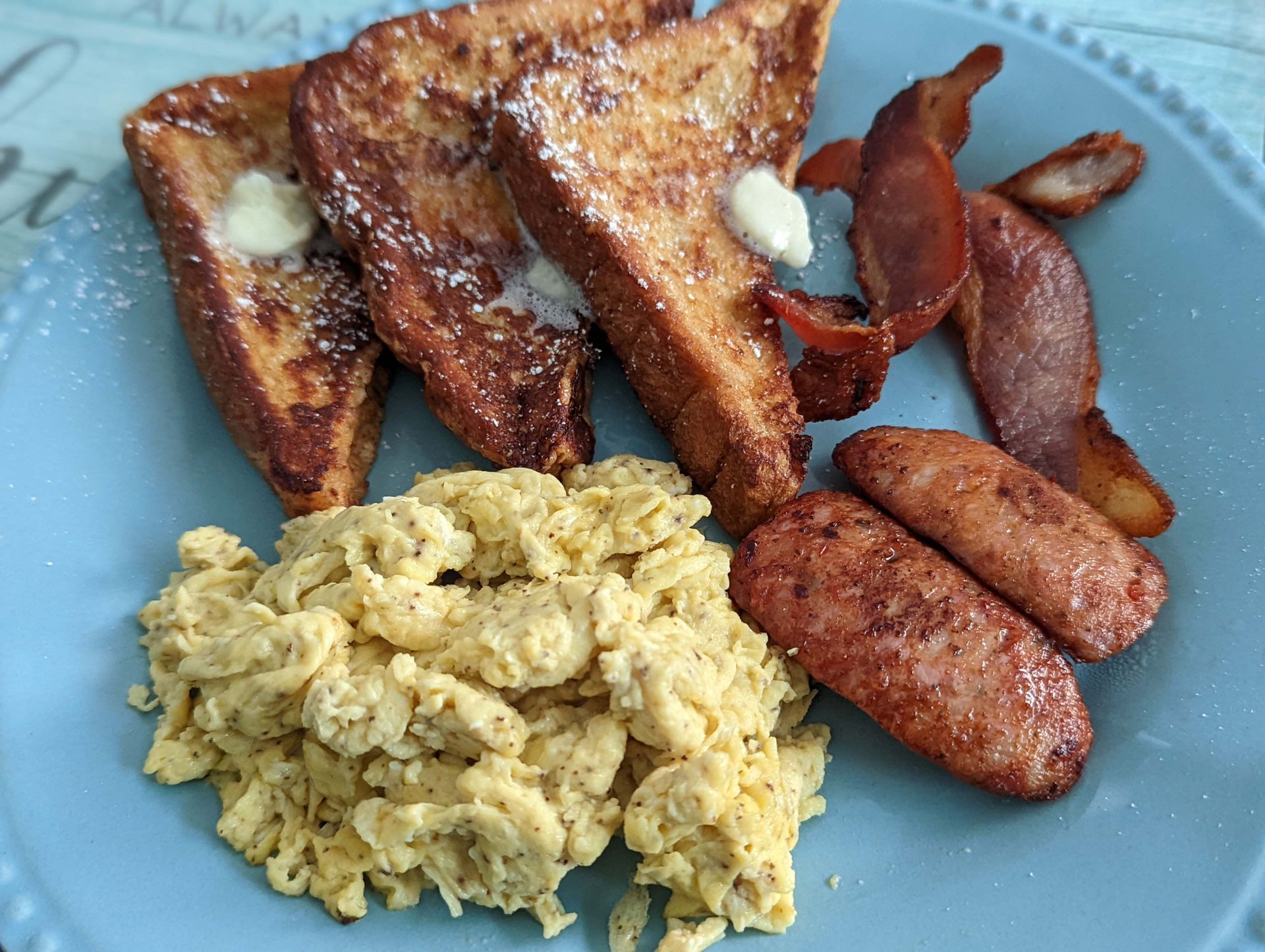 A blue plate topped with french toast , scrambled eggs , bacon and sausage.