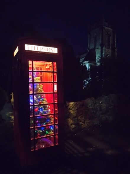 Red British phone booth glowing at night, decorated with a lit Christmas tree inside.