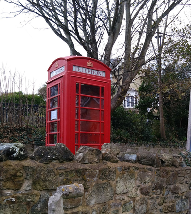 Red British-style phone booth beside a stone wall and bare tree in a garden.