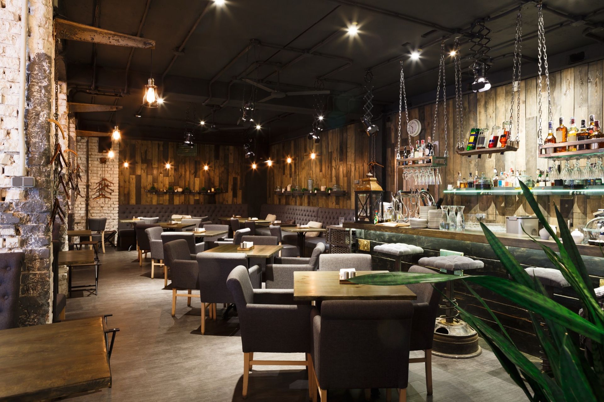 An interior view of a dimly lit restaurant featuring wooden walls, dark-upholstered chairs, and a bar area with hanging lights.