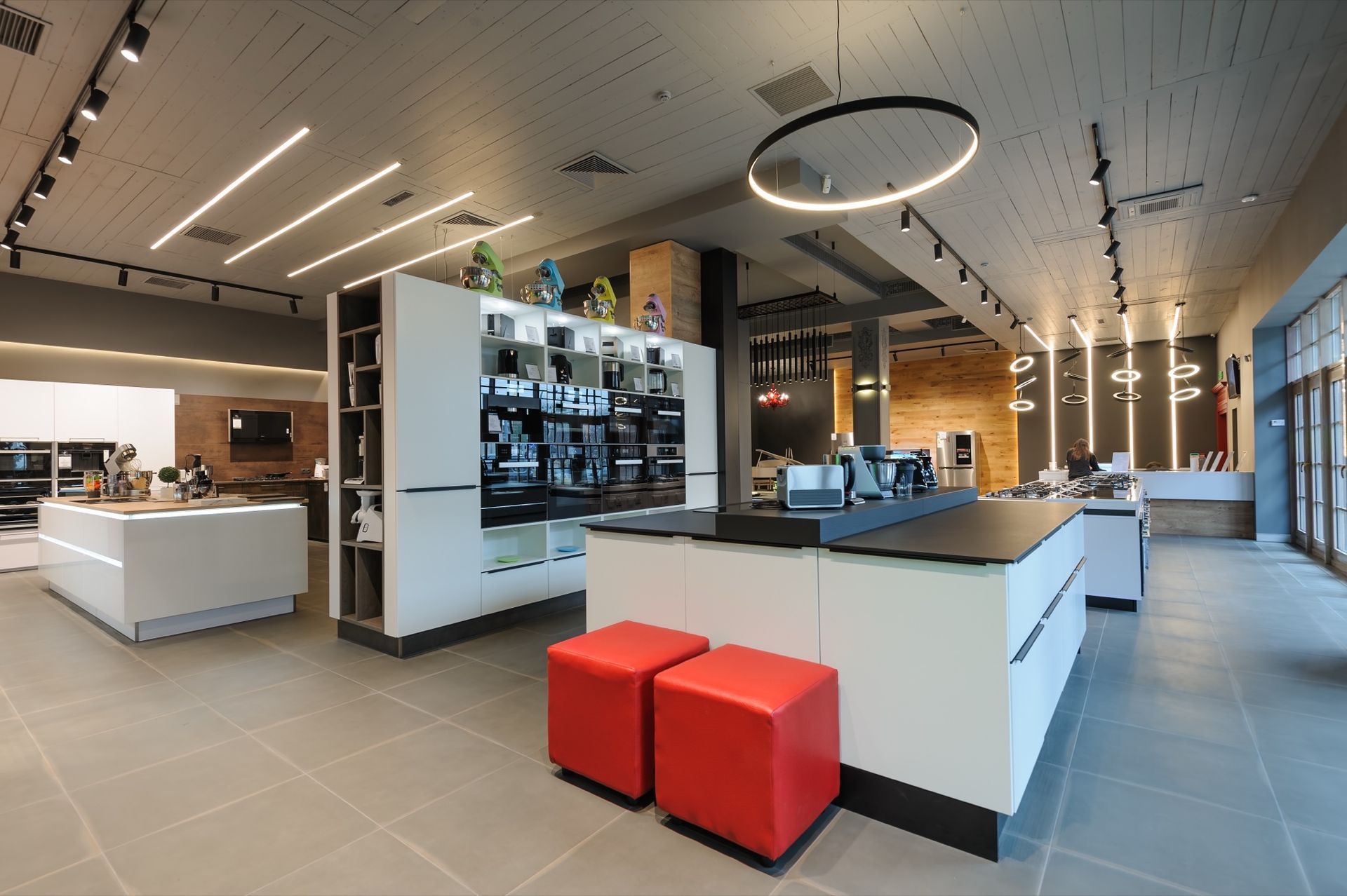 Modern Kitchen Showroom with White Islands, Built-In Appliances, Red Cube Stools, and Minimalist Overhead Lighting.