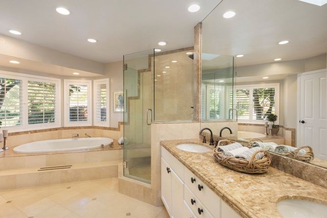 Spacious beige bathroom with soaking tub, glass shower, granite vanity, and large windows.