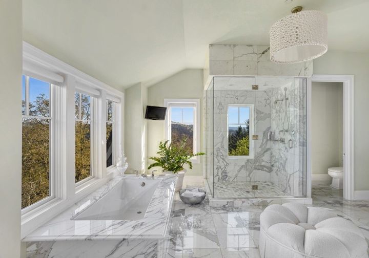 Bright white luxury bathroom with a freestanding tub, marble shower, and large windows overlooking trees