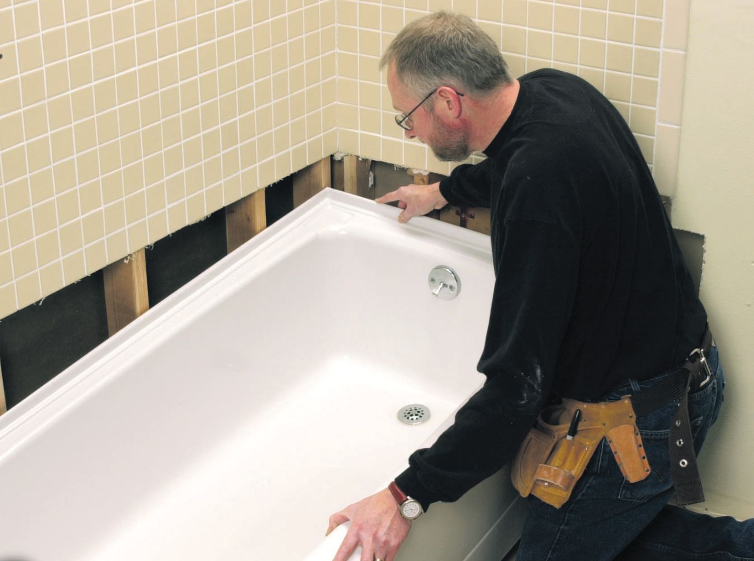 Worker installing a white bathtub in a tiled bathroom, leaning over the rim with tools on his belt.