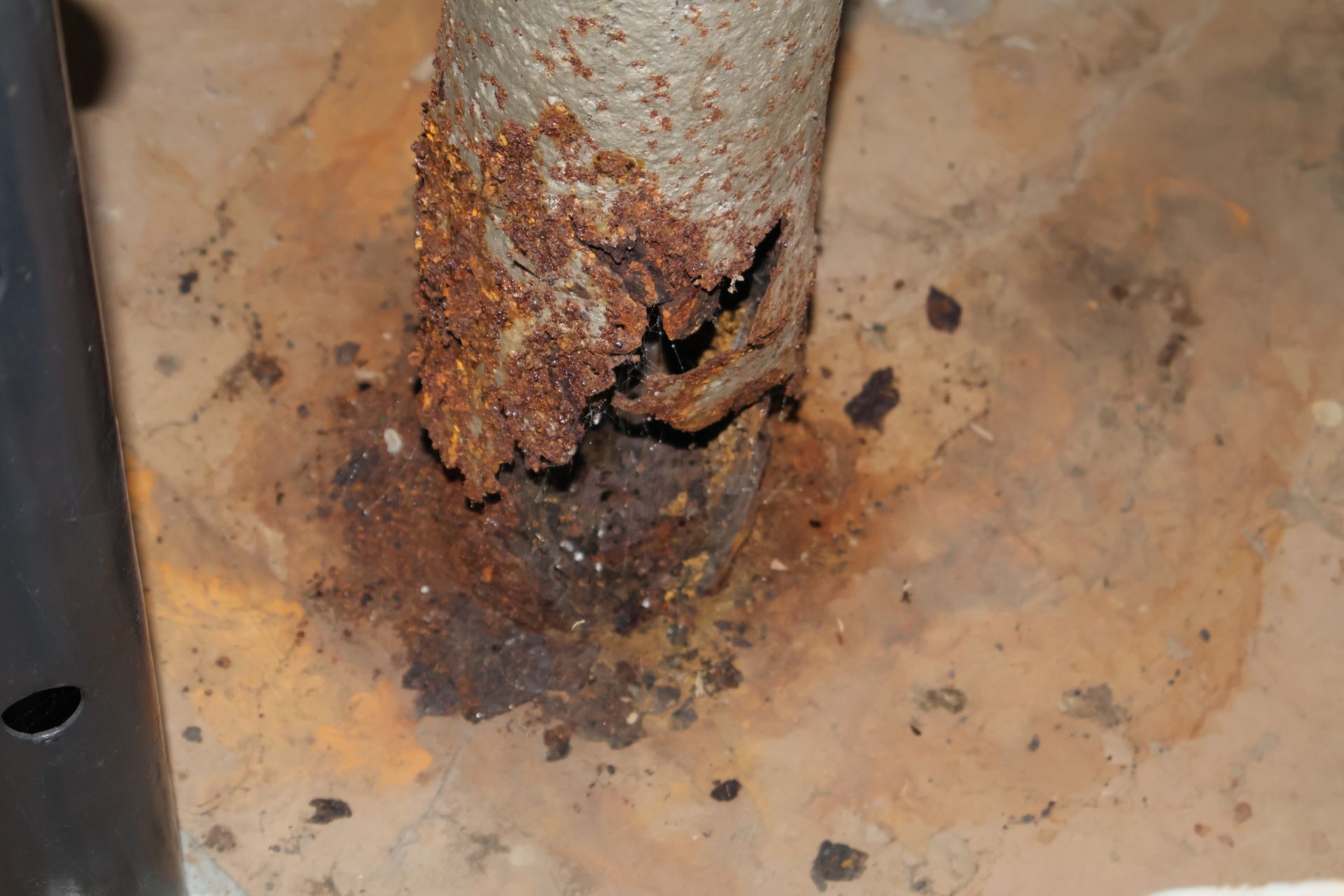 A heavily rusted, corroded metal pipe pierces a concrete floor, with significant structural decay at the base.