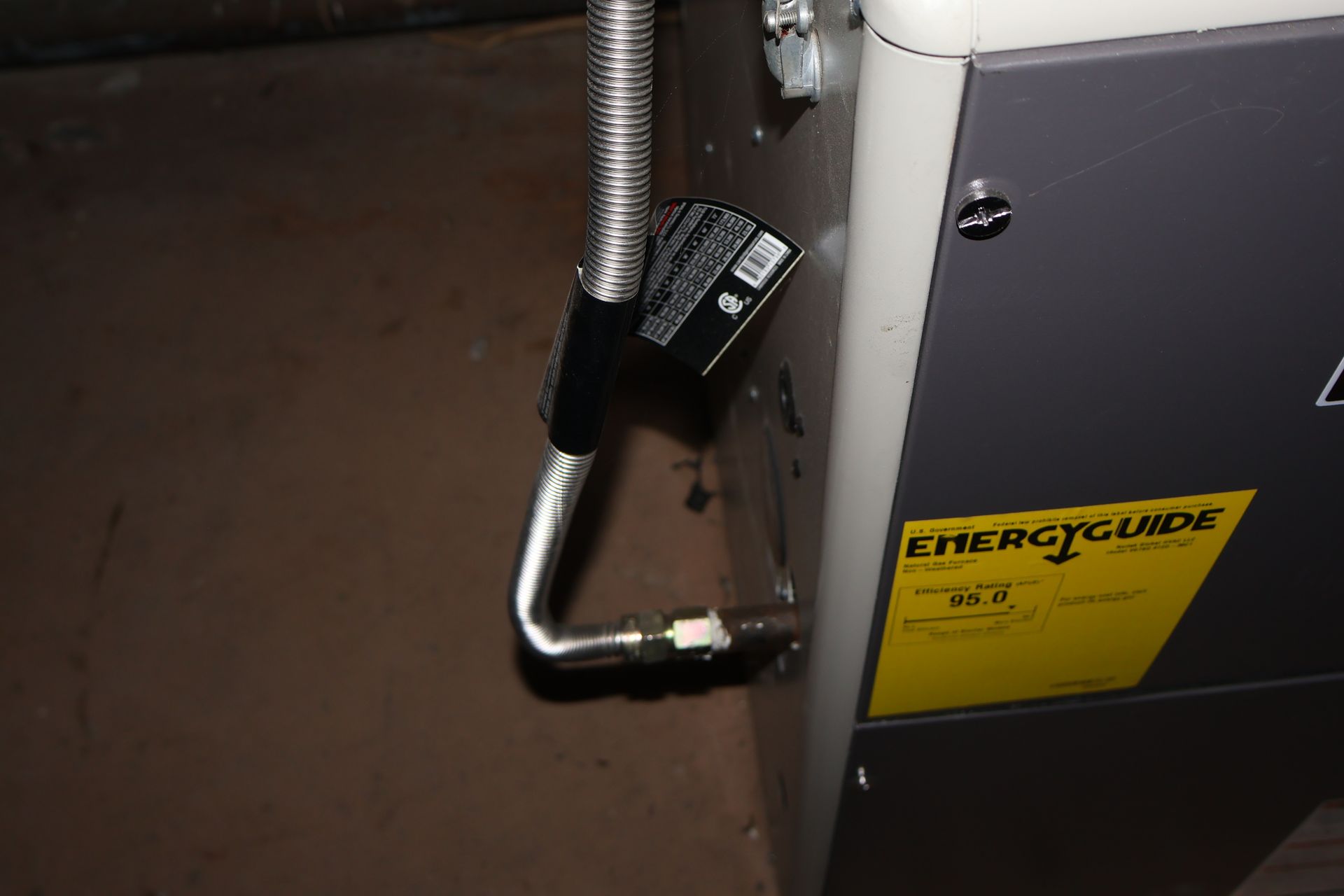 A flexible metal gas line connects to the side of a grey heating unit next to a yellow EnergyGuide label.