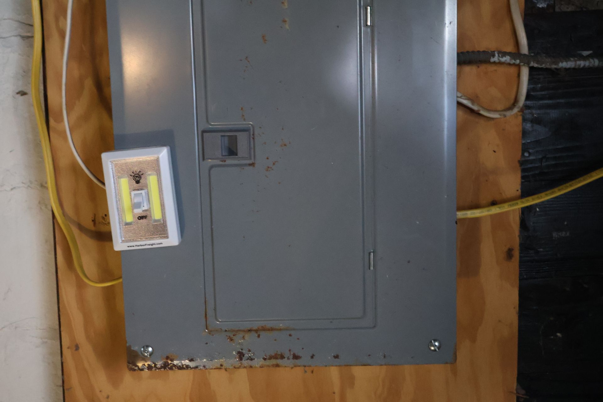 A gray electrical panel mounted on a plywood wall with a small light fixture attached to its front cover.