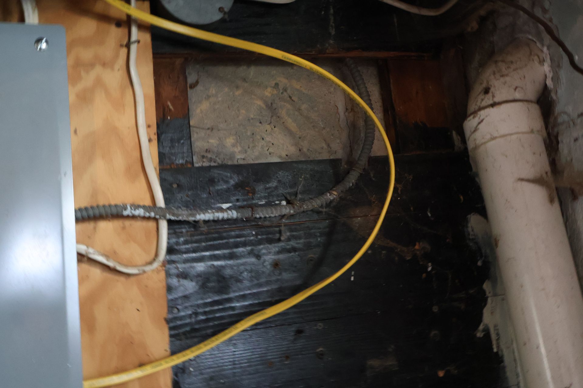 Electrical cables run across dark, stained wooden framing and a vertical white pipe near a grey electrical panel.
