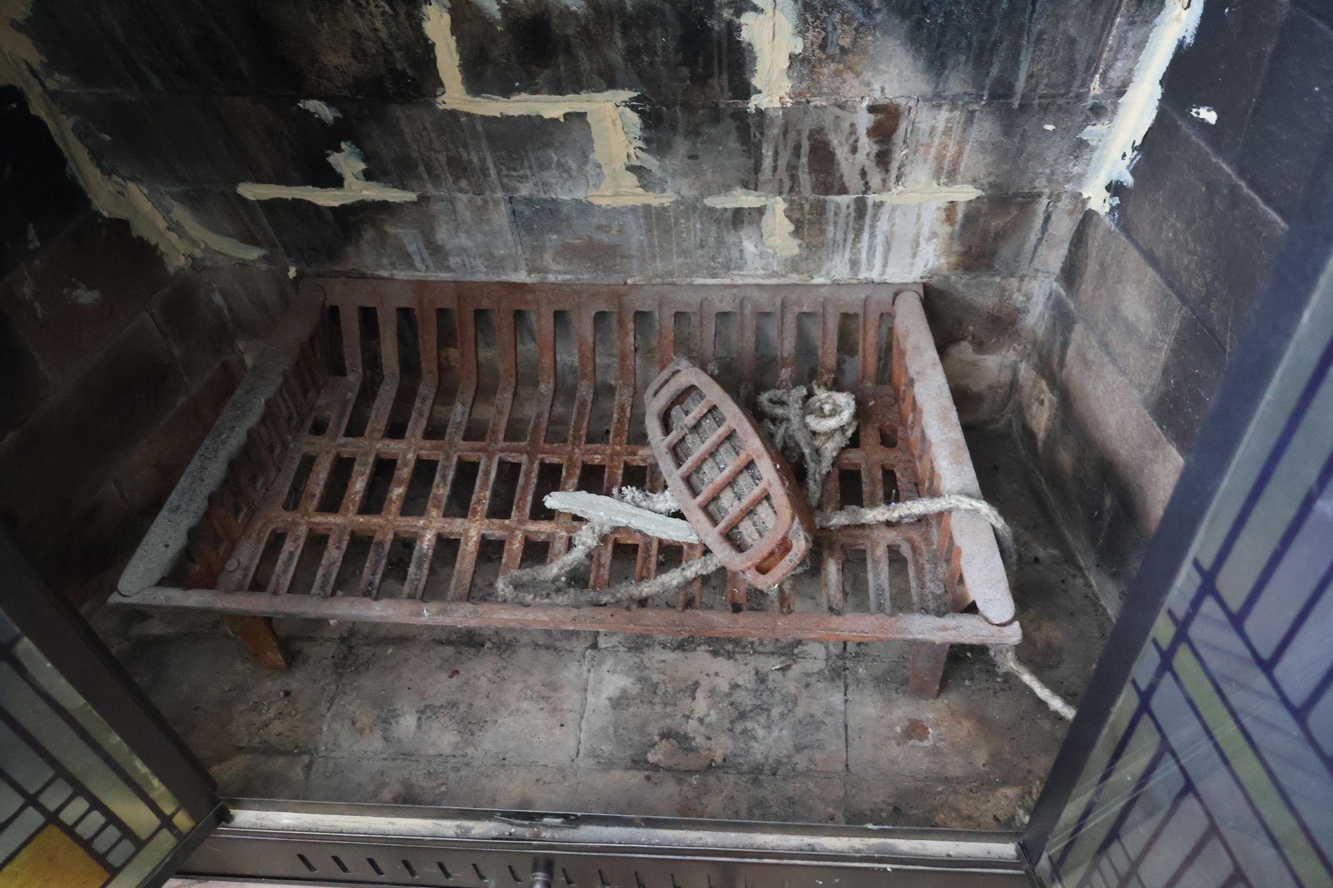 A rusted metal fireplace grate sits inside a hearth with a detached circular heating component and a coiled cord.