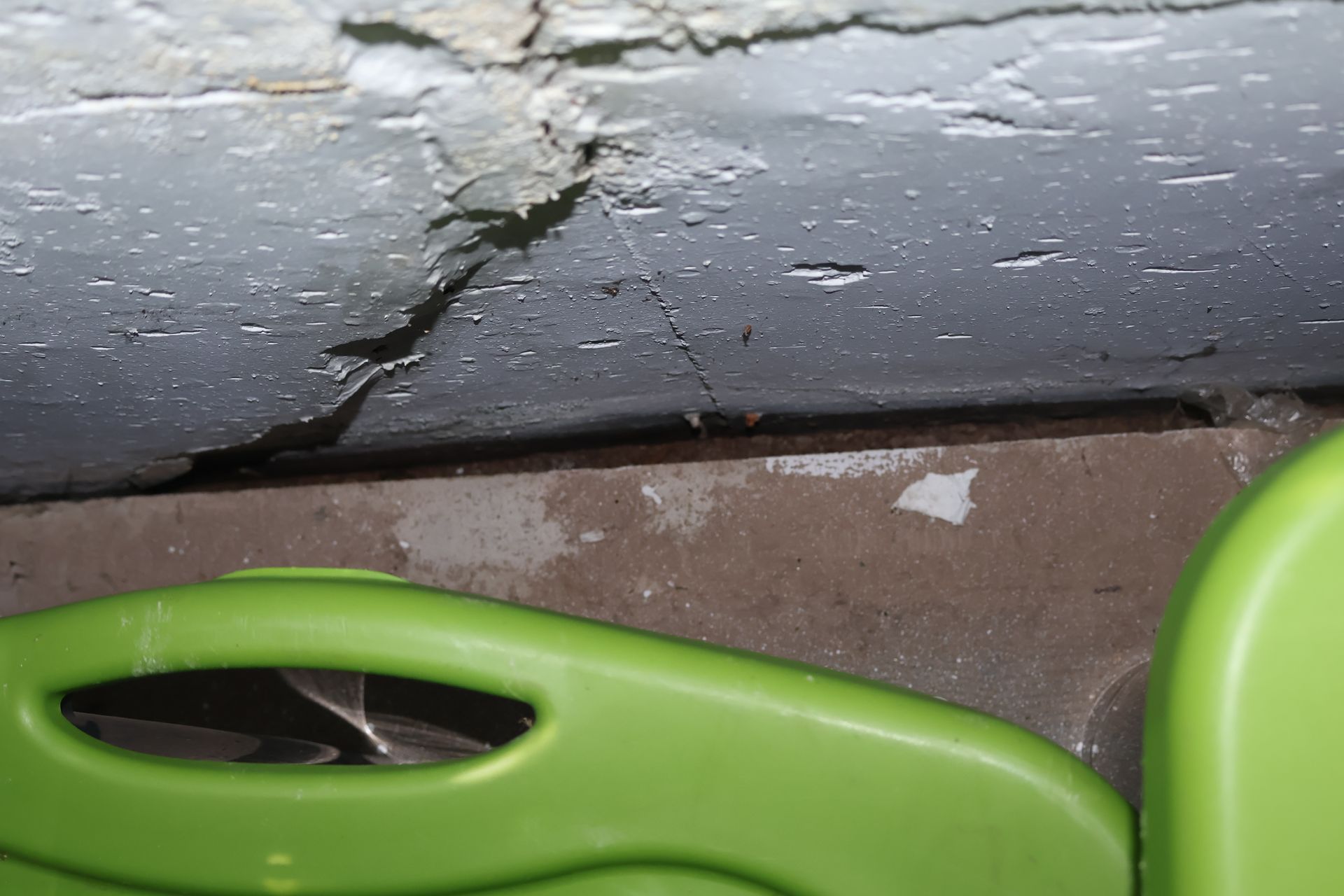 A close-up view of a damaged wall corner with peeling gray paint above a brown surface and a bright green plastic object.