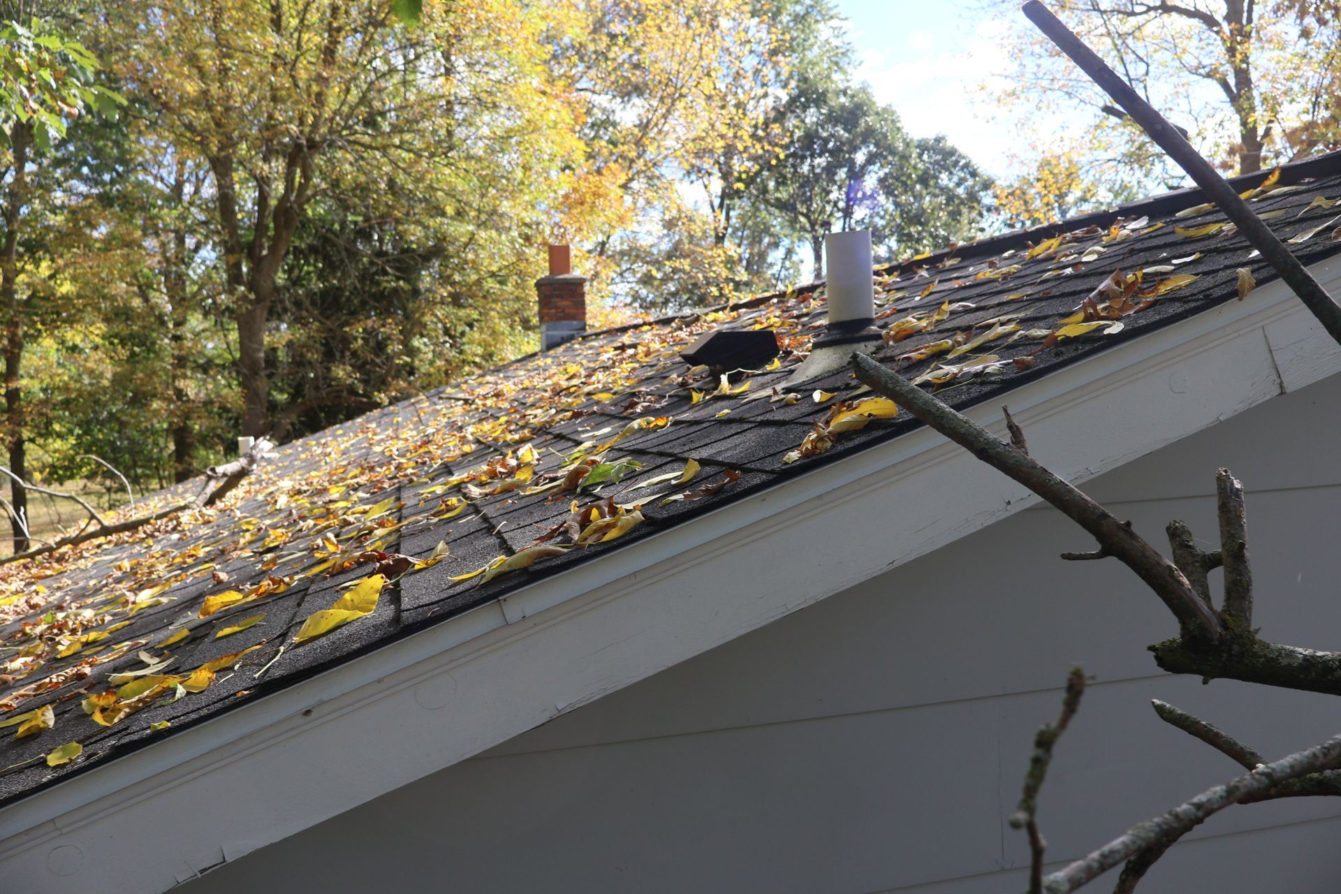 A slanted roof covered in fallen autumn leaves with a brick chimney and a white plumbing vent pipe.