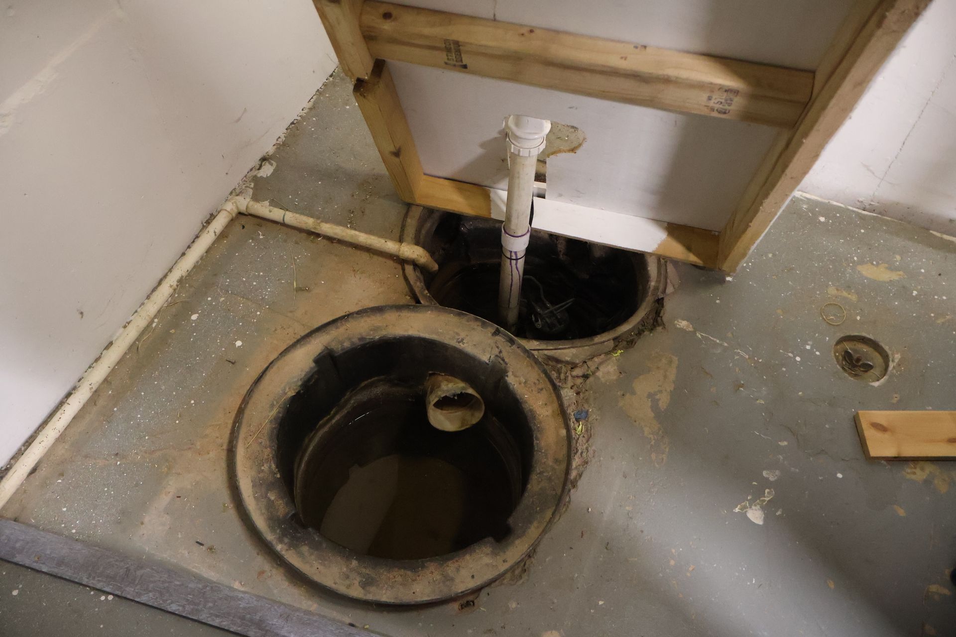 Two open concrete floor drains in a basement near unfinished wood framing and plumbing pipes.