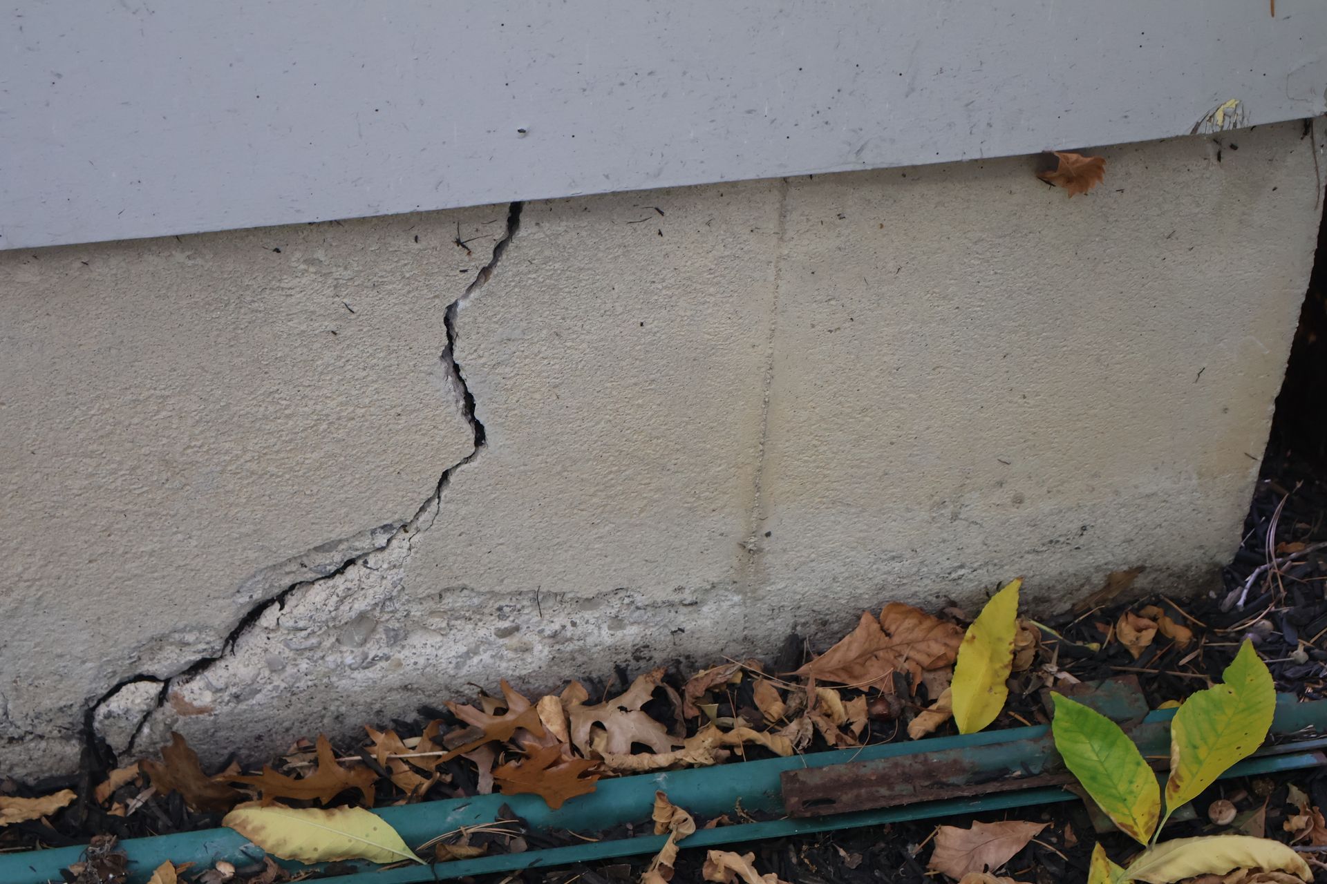 A jagged diagonal crack runs through a light-colored concrete foundation wall above a ground area with fallen leaves.