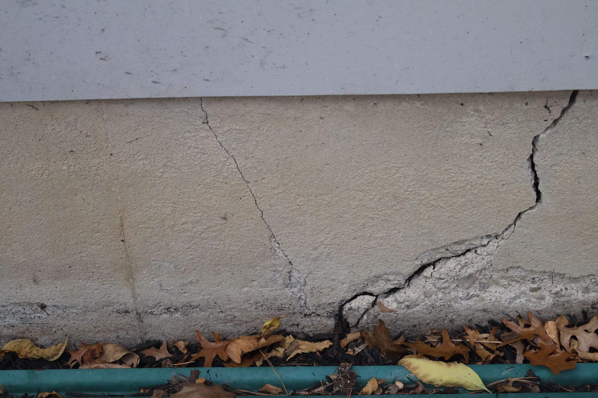 A close-up of a house foundation with a diagonal crack extending from the ground upward toward the siding.