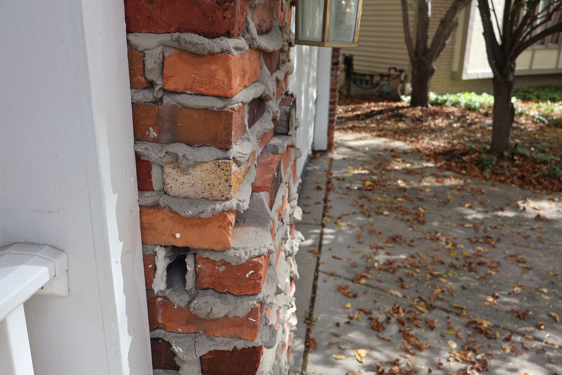 A side view of a brick column with visible gaps and rough, uneven mortar, next to a white post and a sidewalk.