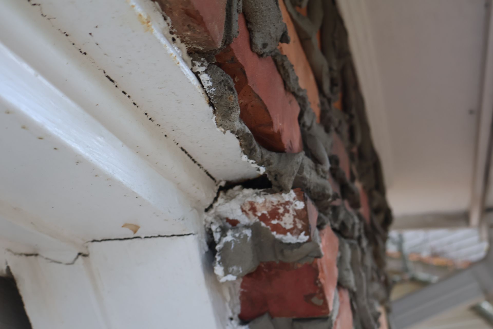 A close-up of a damaged exterior door frame where white wood trim meets red brick wall with visible gaps and mortar.