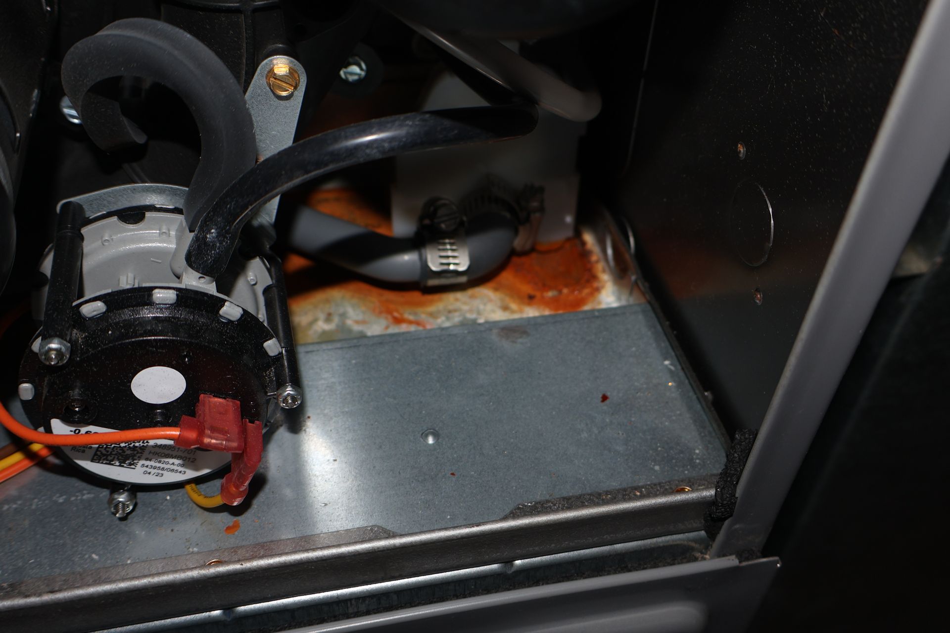 Inside a furnace cabinet, orange rust stains appear on the metal base near a drainage hose and pressure switch.