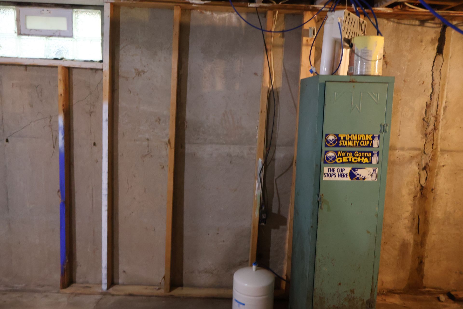 A basement wall with exposed wood studs, a small rectangular window, and a green metal locker with stickers.