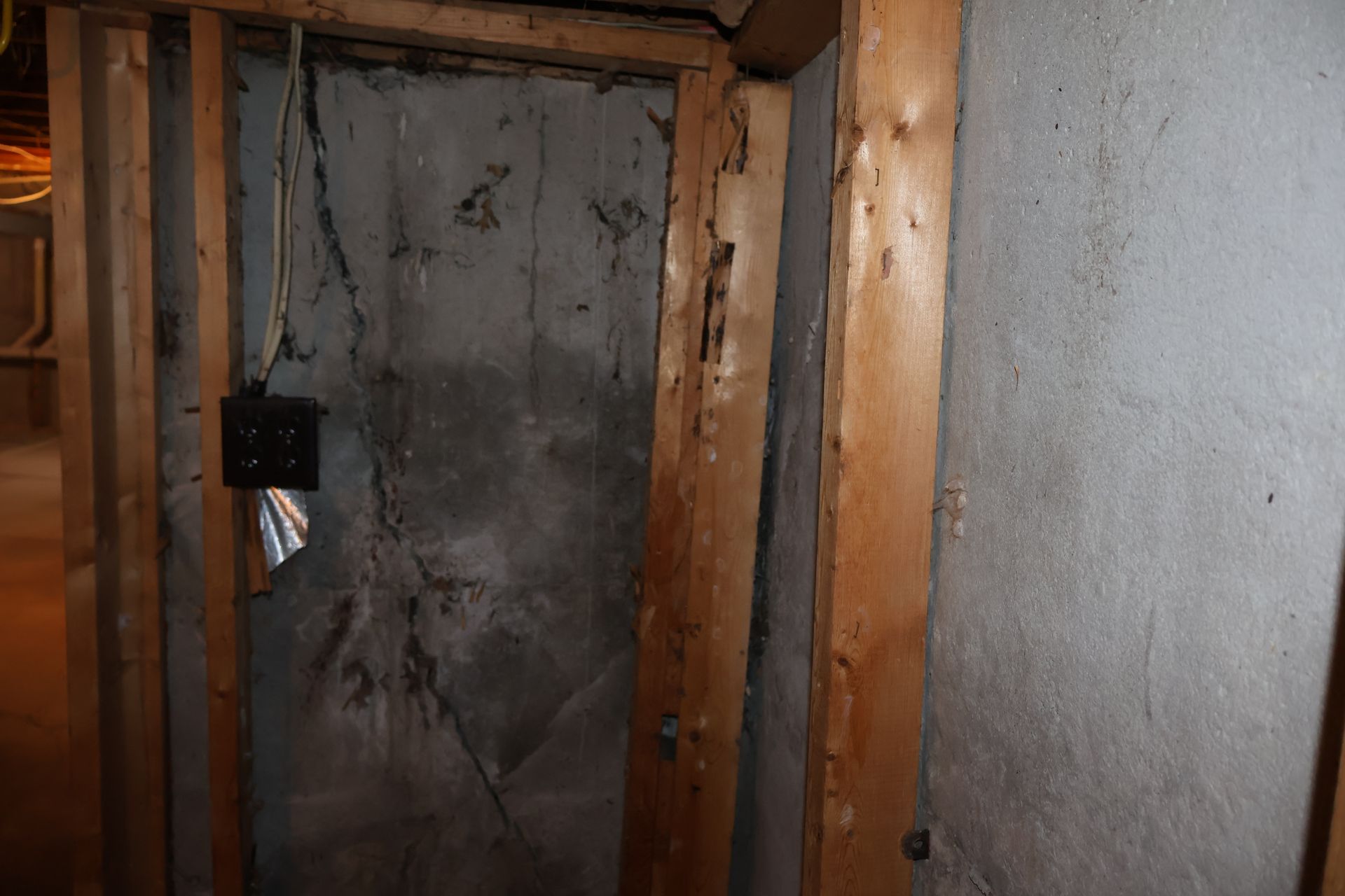 A corner of an unfinished basement shows a dark, cracked concrete wall framed by wooden studs and an electrical box.