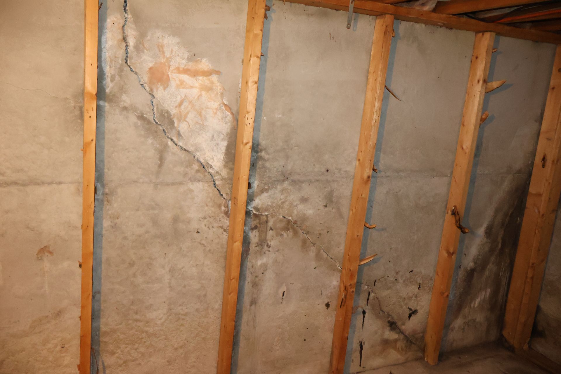 A basement concrete wall with prominent diagonal cracks and water staining, partially framed by wooden studs.