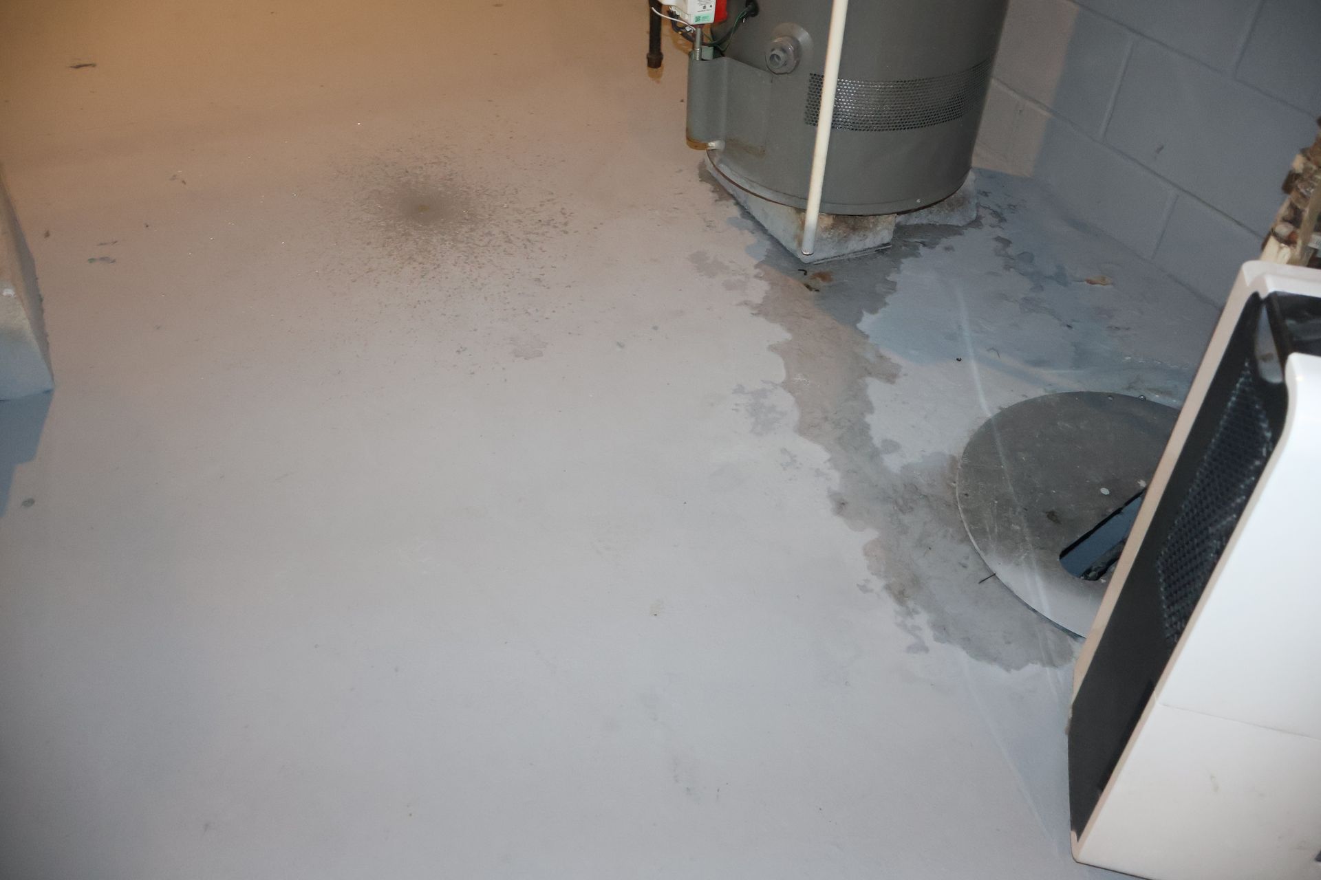 A gray basement floor shows a water stain leading from a water heater to a circular floor drain.